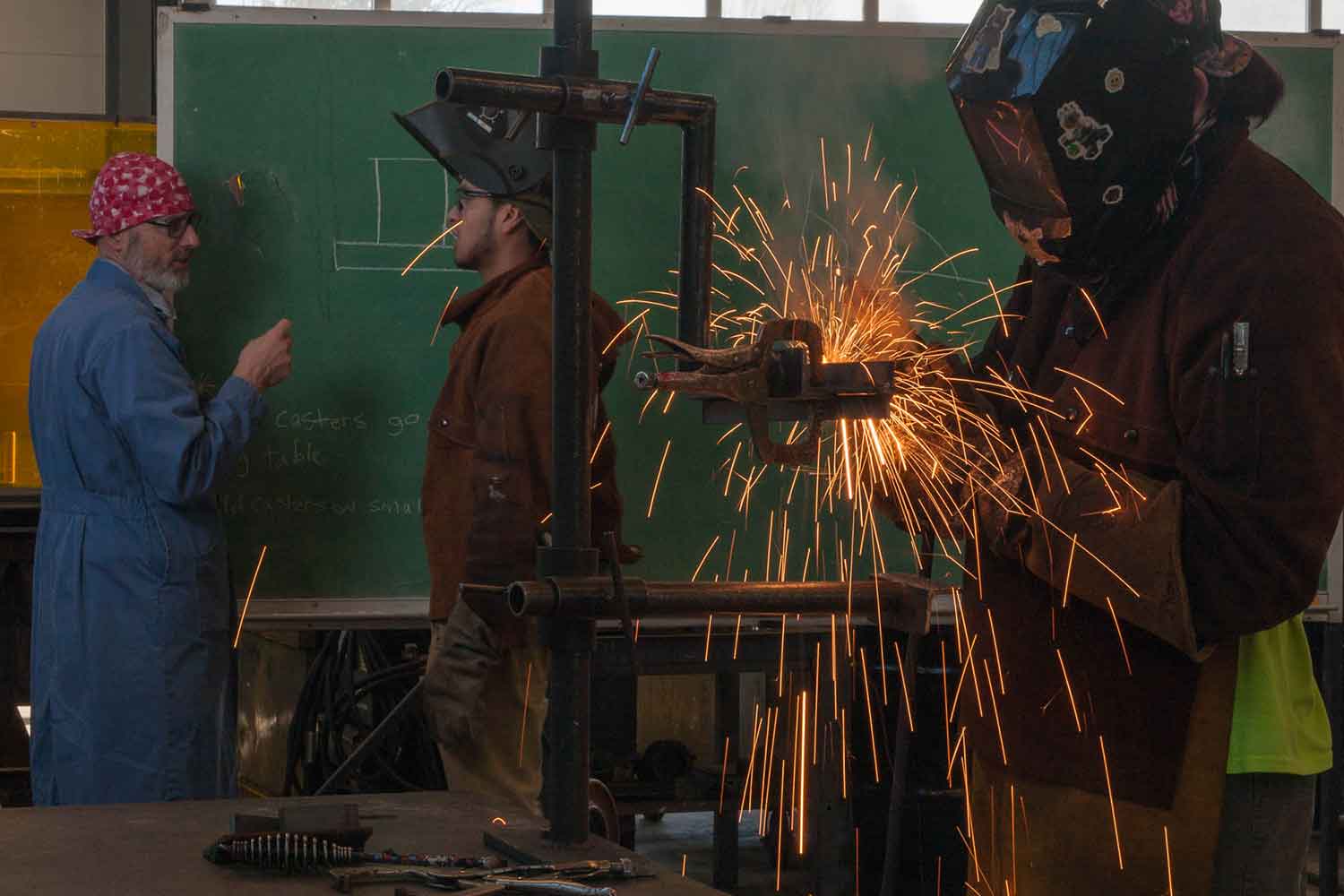 A student welds while an instructor teaches another student in front of a blackboard